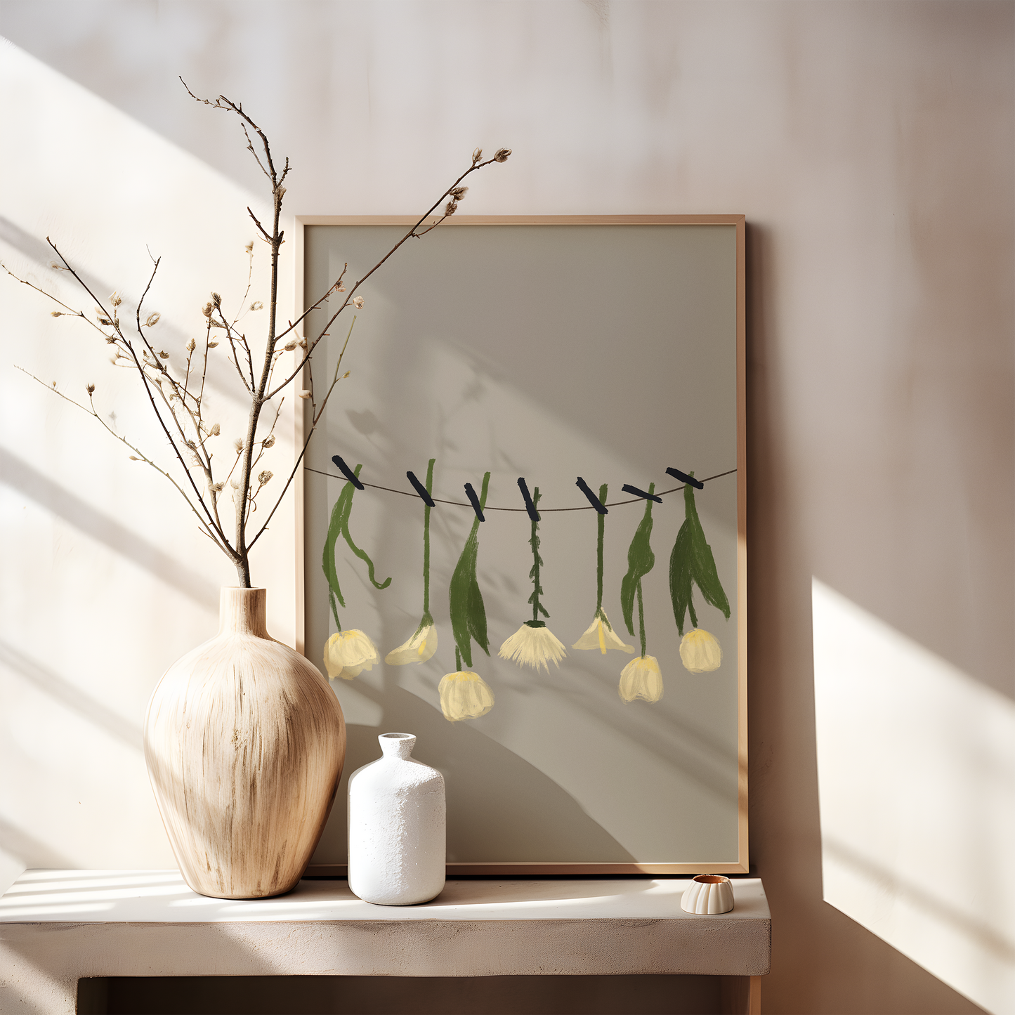 A framed digital print featuring stylized white flowers suspended on a line, placed on a wooden console table with simple ceramic vases and dry branches, casting soft shadows in natural light