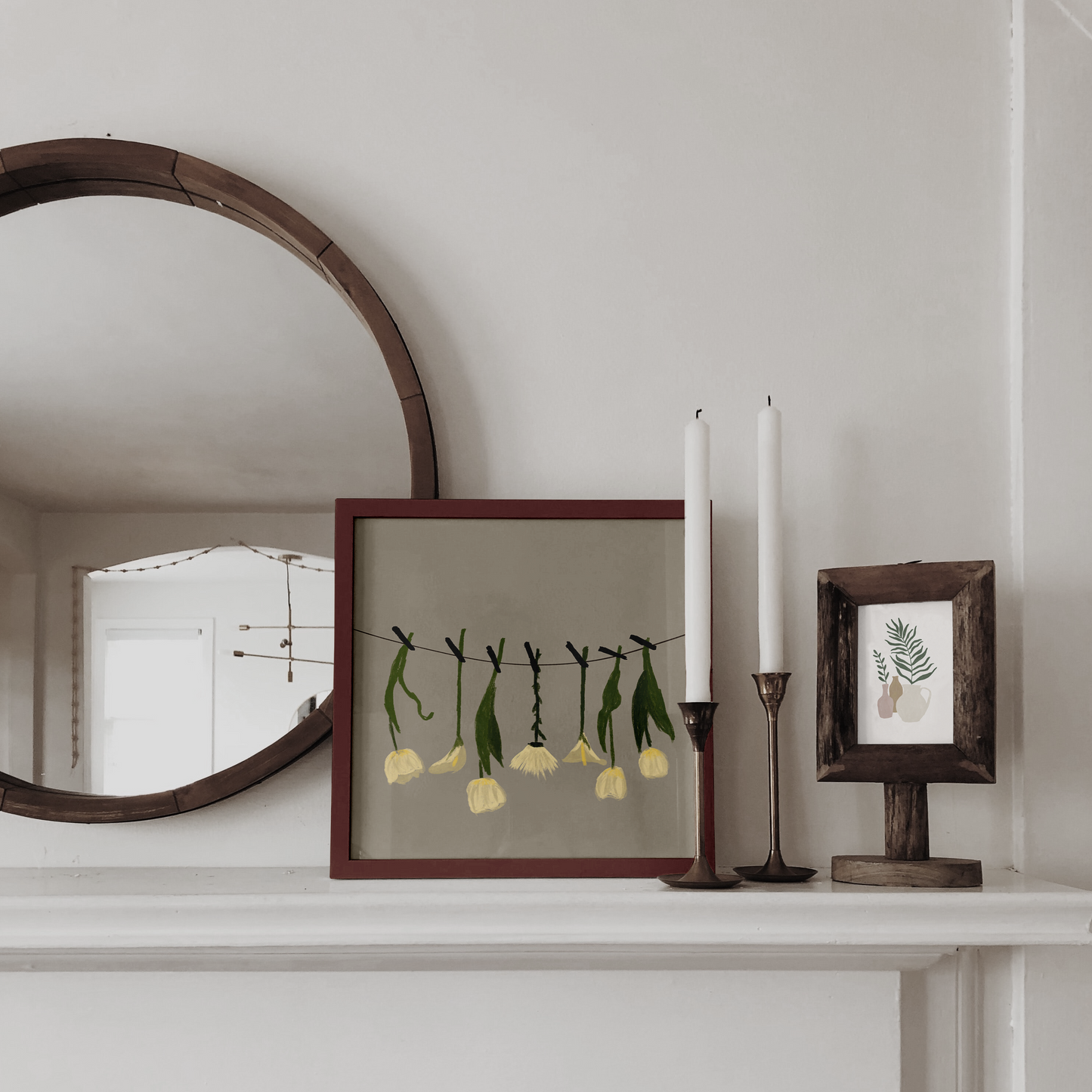 A rustic-chic digital painting of white flowers on a string, encased in a red-brown frame on a mantelpiece, creating a charming vintage feel with candles and a wooden mirror.